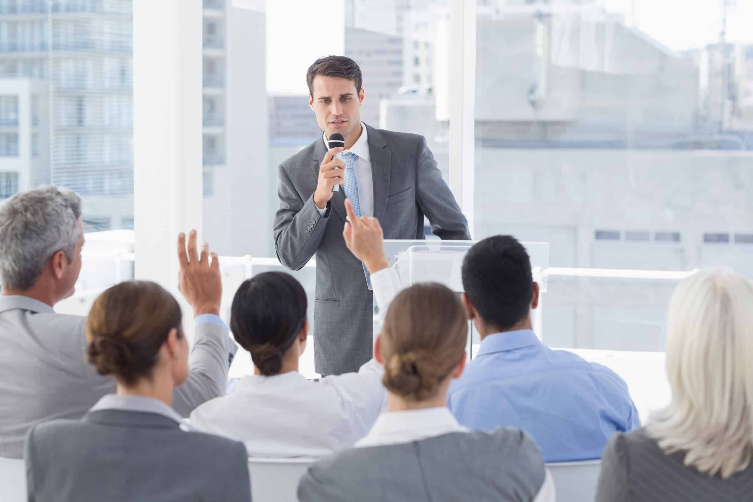 Business man stands a lectern in a meeting room, taking questions from a group of employees seating in front of him.