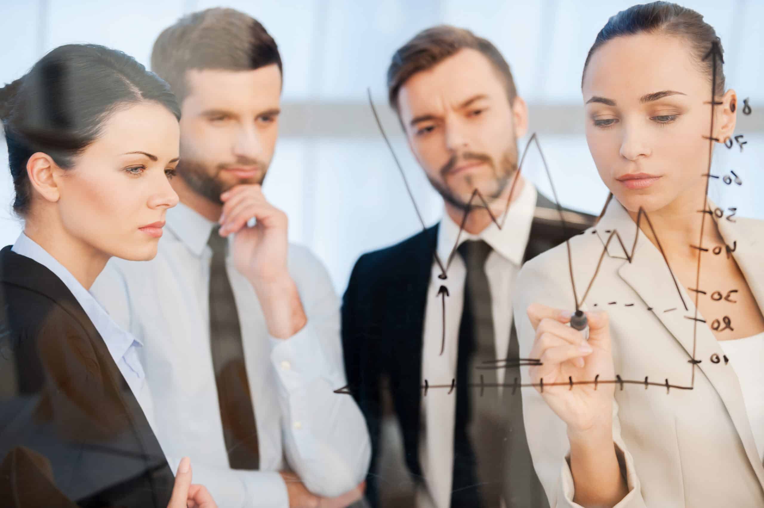 Business woman draws a line graph on a clear screen, while colleagues watch.