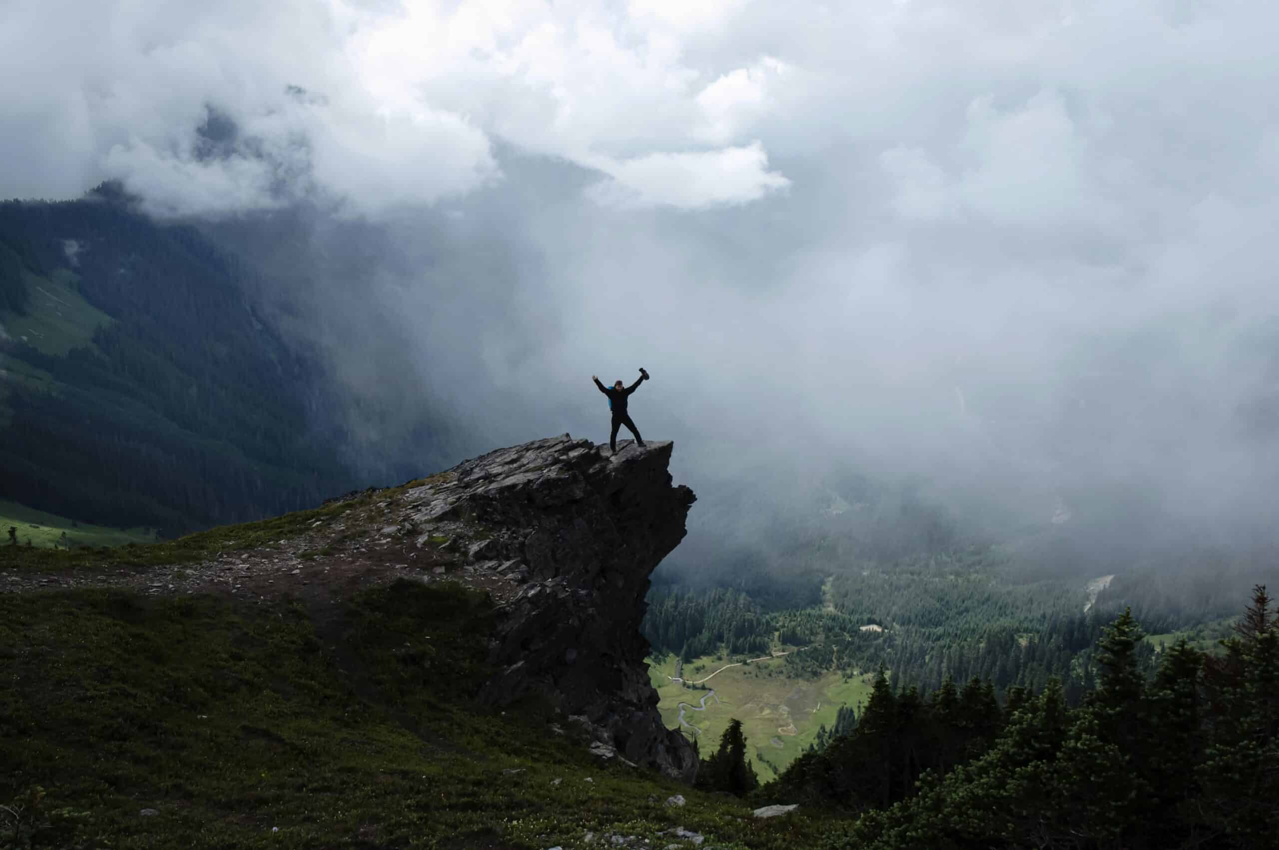 Climber at rocky summit in the clouds above a valley raises arms triumphantly