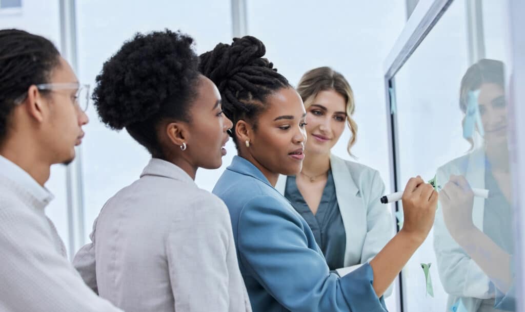 Business woman writes on a white board, while colleagues stand behind her watching.
