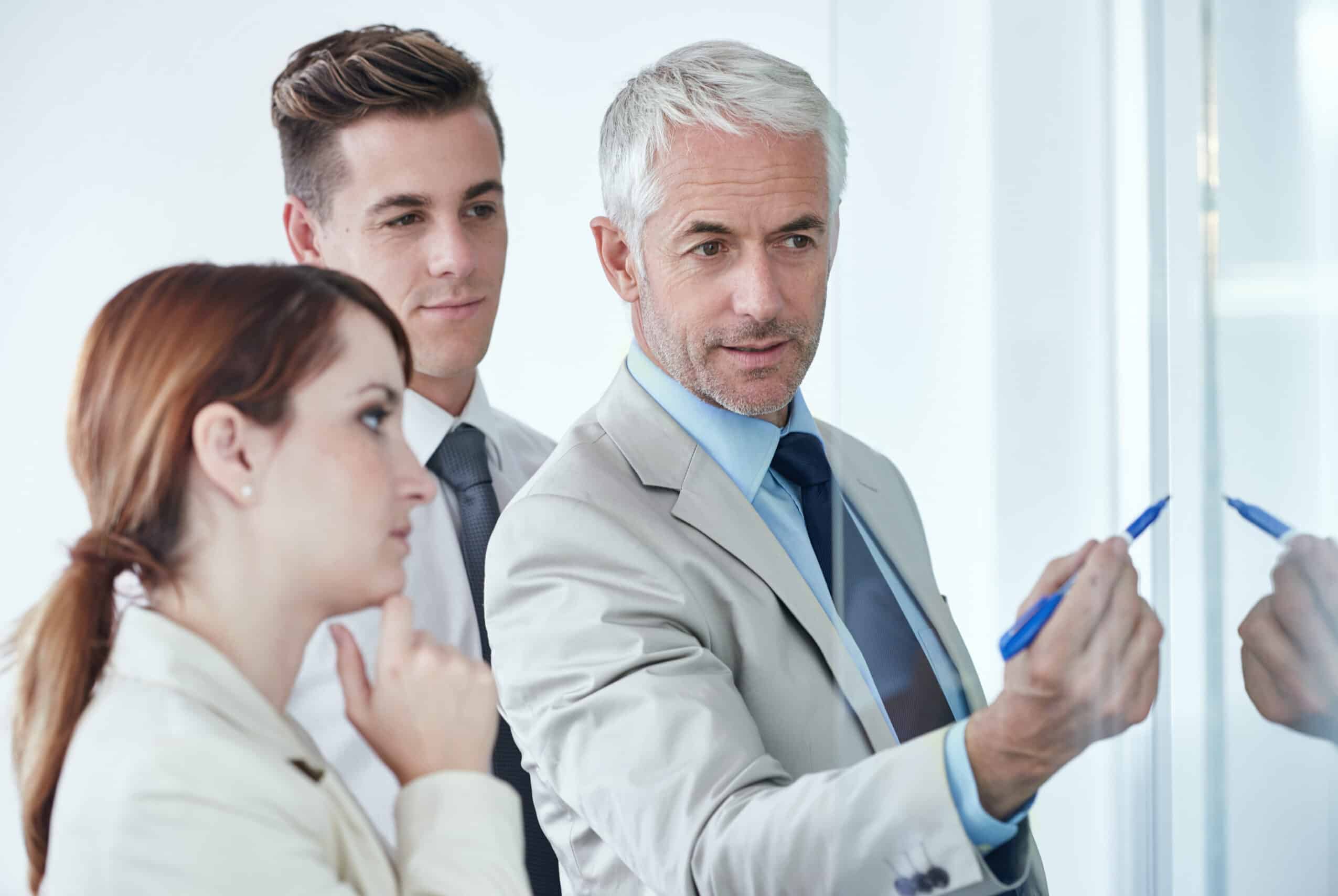 Business man writes concepts on a marker board while associates stand behind him watching.